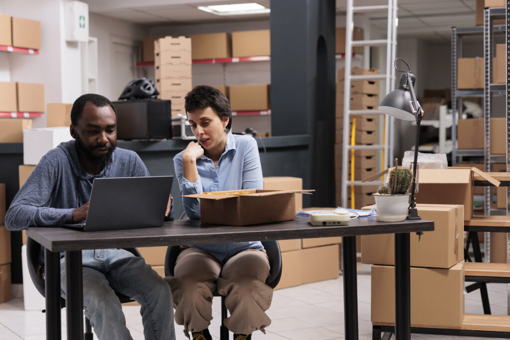 Employees looking at cargo stock on laptop after finishing packing trendy clothes in carton boxes